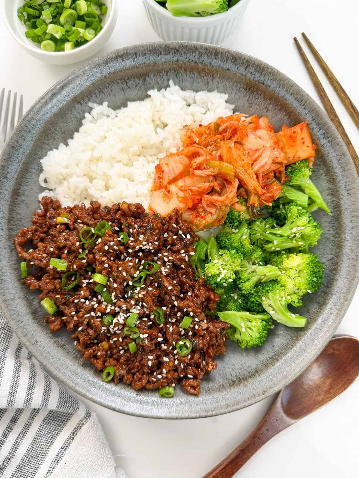 A gray bowl with white rice, seasoned Sticky Gochujang Ground Beef Bowls topped with sesame seeds and green onions, kimchi, and broccoli. Chopsticks, a spoon, and a striped napkin are nearby.
