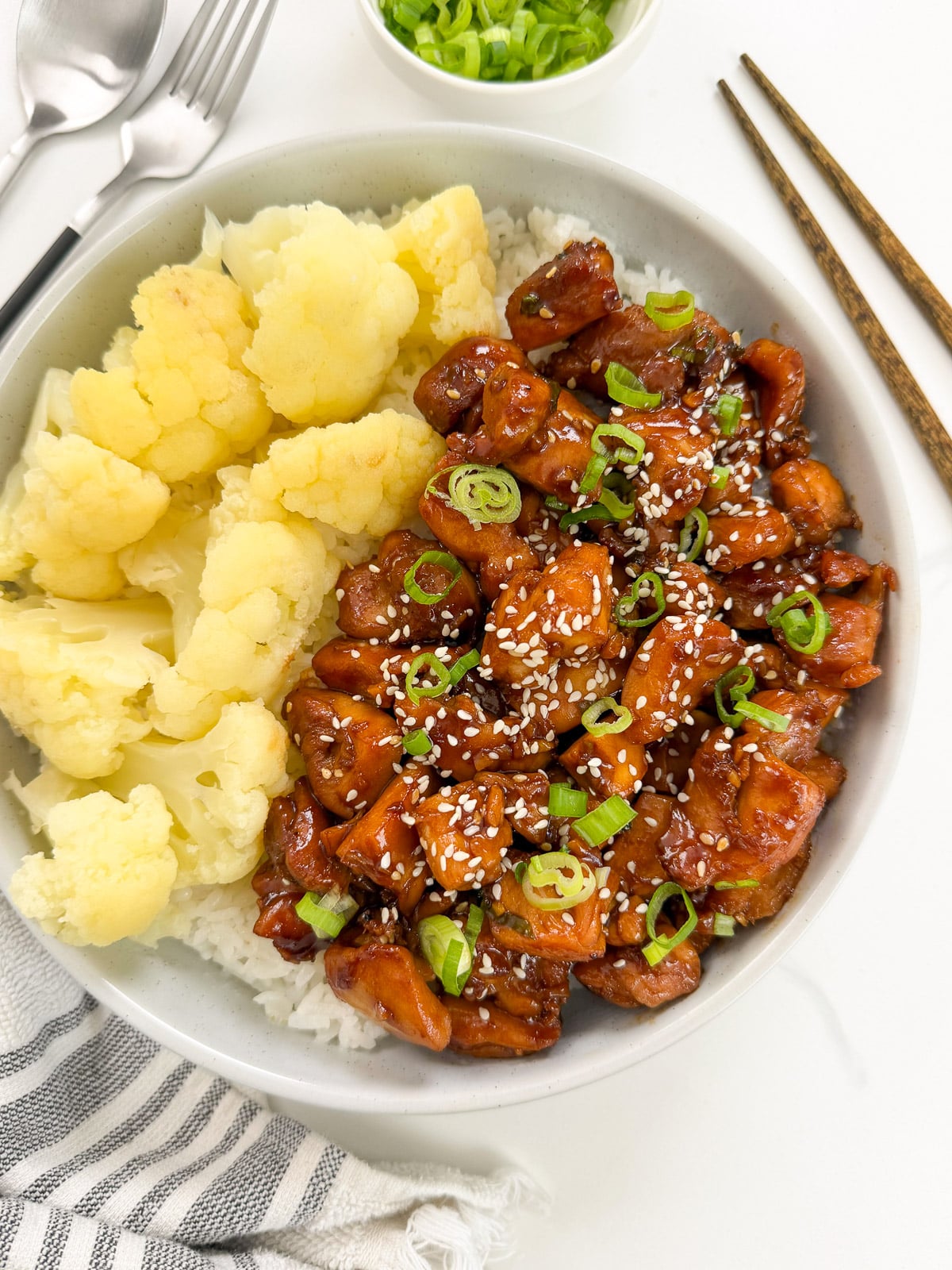A bowl of white rice topped with cooked cauliflower and Sweet and Sticky Hoisin Chicken pieces, garnished with sesame seeds and chopped green onions. Chopsticks, spoon, and fork are beside the bowl.
