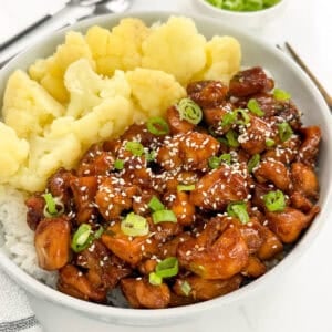 A bowl of rice topped with sweet and sticky hoisin chicken pieces, sliced green onions, and sesame seeds, served with steamed cauliflower florets.