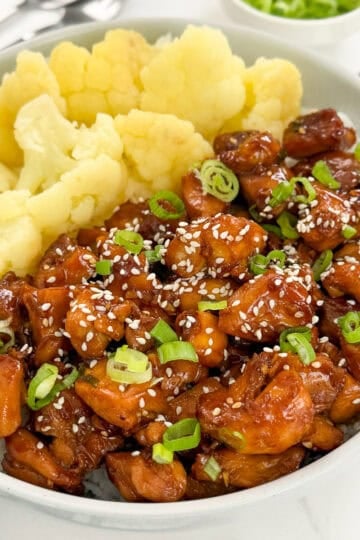 A bowl of rice topped with sweet and sticky hoisin chicken pieces, sliced green onions, and sesame seeds, served with steamed cauliflower florets.