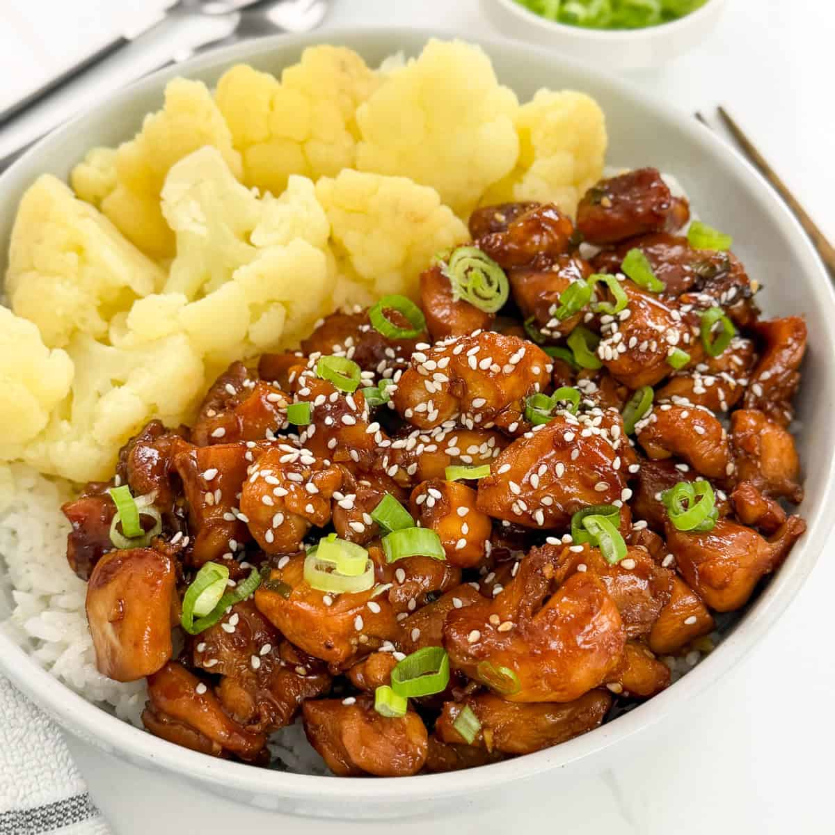 A bowl of rice topped with sweet and sticky hoisin chicken pieces, sliced green onions, and sesame seeds, served with steamed cauliflower florets.