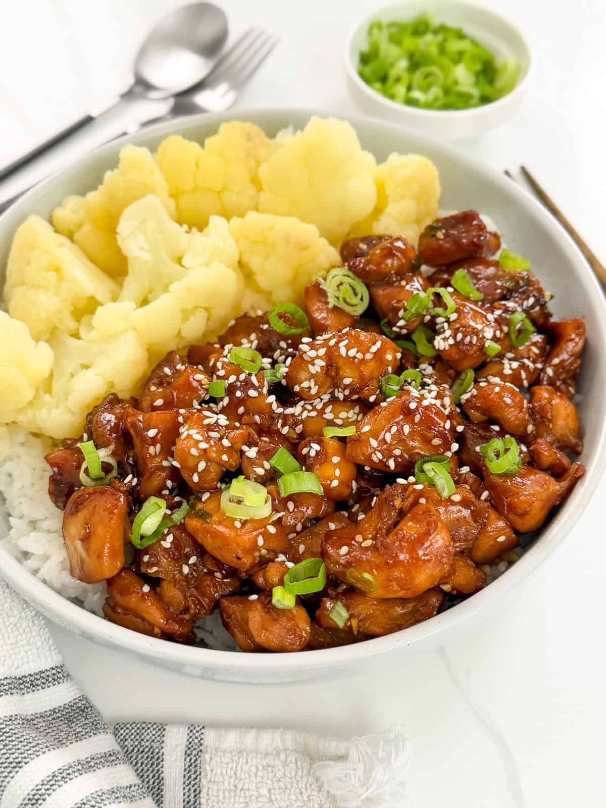 A bowl of rice topped with sweet and sticky Hoisin chicken pieces, sprinkled with sesame seeds and chopped green onions, served with steamed cauliflower. A small bowl of green onions and utensils are in the background.