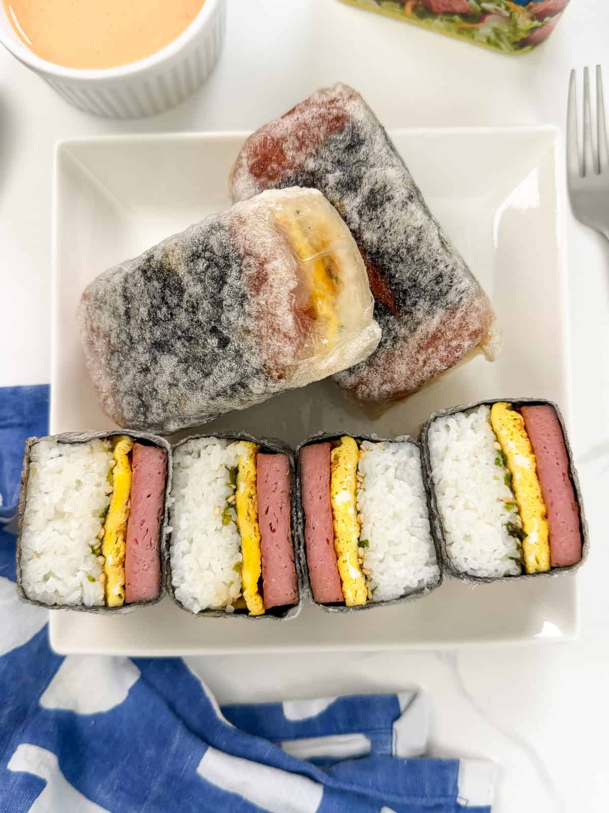 A plate with two whole and four sliced Teriyaki Spam musubi, wrapped in seaweed, rice, spam, and egg. A small bowl of sauce and a blue and white napkin are nearby on a white surface.