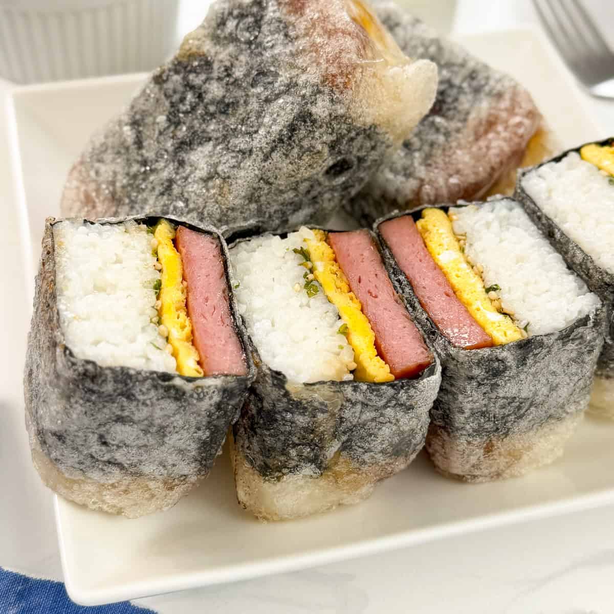 A plate of Teriyaki Spam Rice Paper Musubi, featuring layers of rice, egg, and pink meat wrapped in seaweed and crispy rice paper tempura batter on the outside.