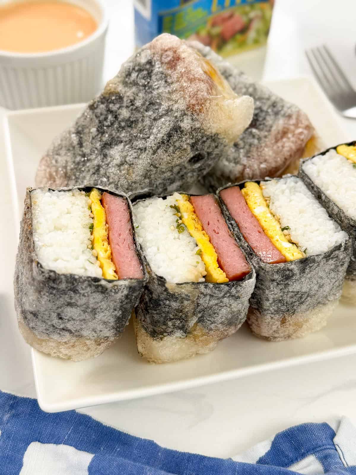 Sliced Teriyaki Spam musubi with egg and rice, wrapped in seaweed, arranged on a white plate. A dipping sauce, a drink carton, and utensils are in the background.