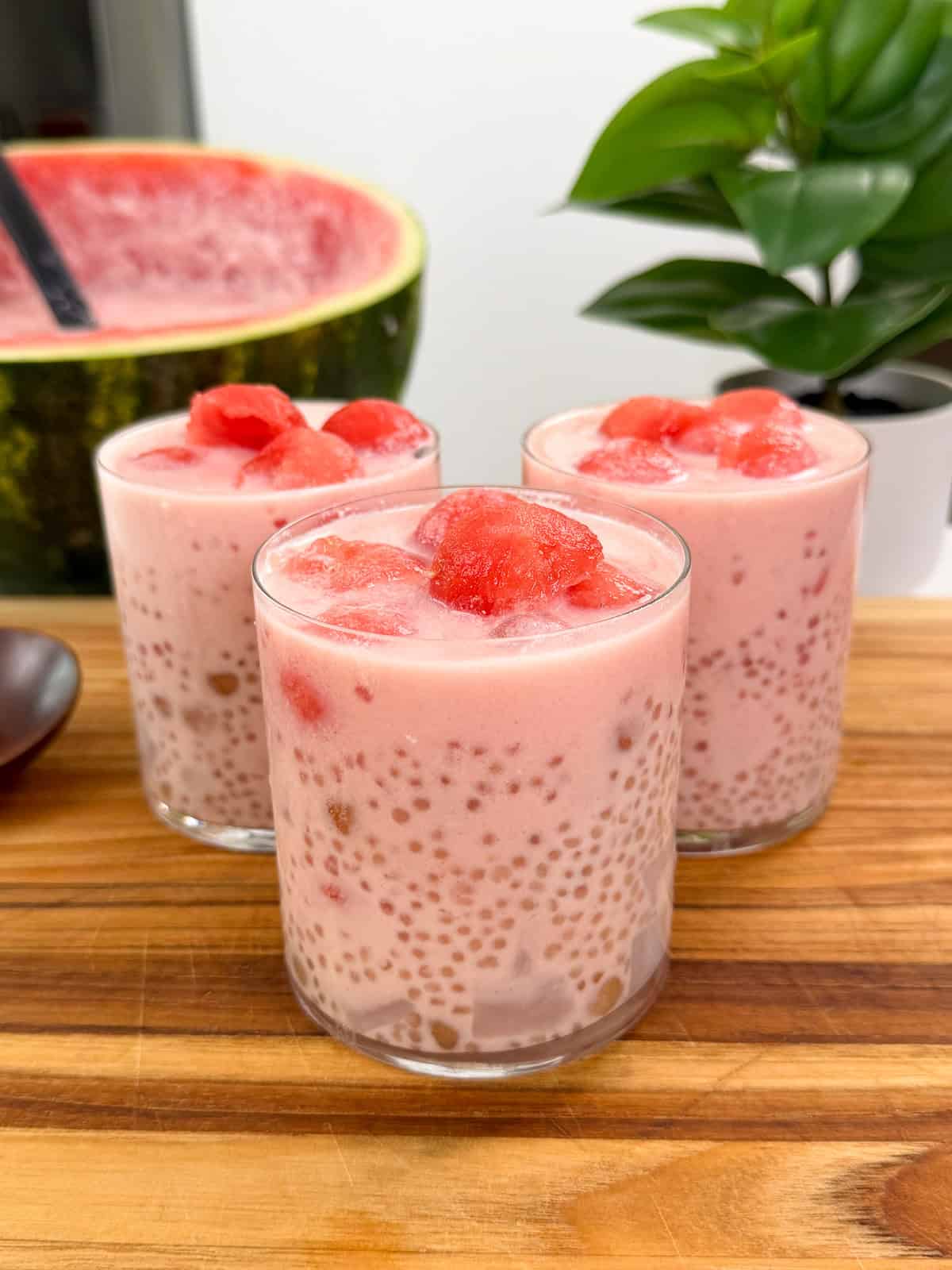 Three glasses of pink watermelon sago pudding topped with juicy watermelon chunks sit on a wooden surface, with a large watermelon and a green potted plant visible in the background.