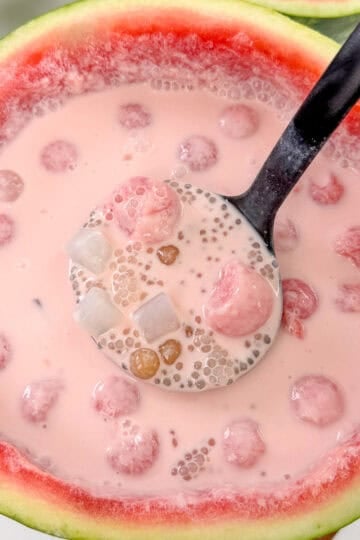 A ladle scoops a pink, milky dessert with tapioca pearls, fruit balls, and jelly cubes—classic ingredients of Watermelon Sago—served in a hollowed-out watermelon bowl.