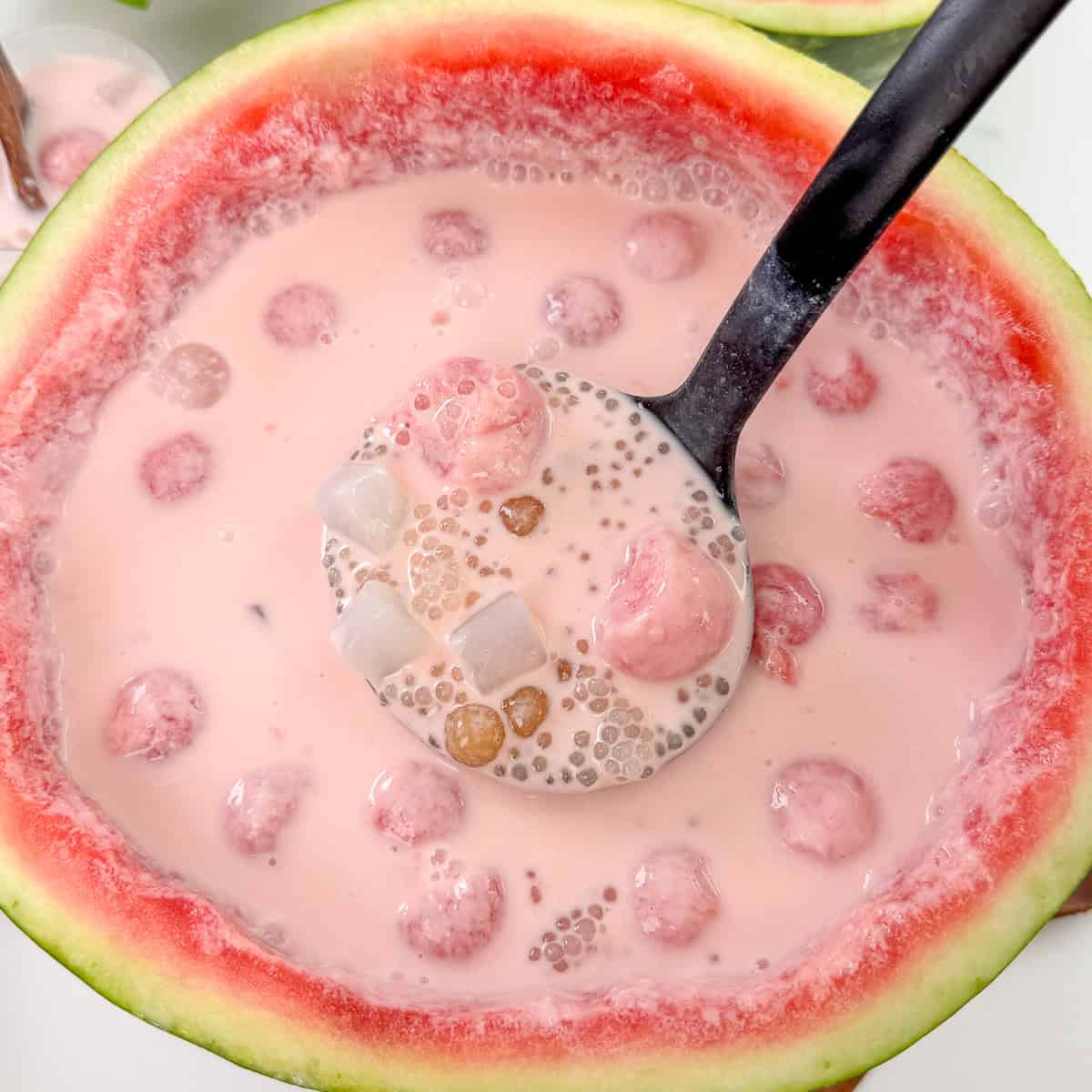 A ladle scoops a pink, milky dessert with tapioca pearls, fruit balls, and jelly cubes—classic ingredients of Watermelon Sago—served in a hollowed-out watermelon bowl.
