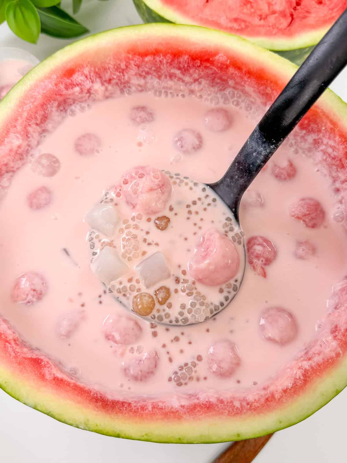 A refreshing watermelon bowl filled with pink milk-based dessert, tapioca pearls, and jelly cubes—perfect for summer. A black ladle scoops up the luscious mixture of watermelon sago, with juicy watermelon flesh visible around the bowl’s rim.
