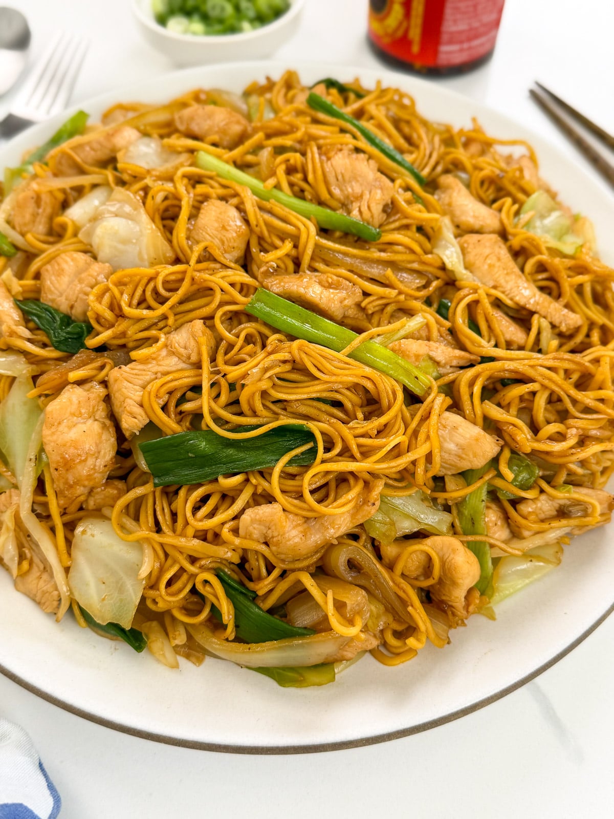 A plate filled with Chicken Chow Mein—stir-fried noodles, tender chicken pieces, bok choy, cabbage, and green onions—is served on a white dish with a fork, knife, and sauces visible in the background.