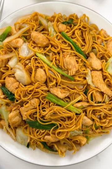 A plate of Chicken Chow Mein featuring stir-fried noodles with tender chicken pieces, cabbage, and green onions. A fork, spoon, and chopsticks are placed nearby on a white surface.