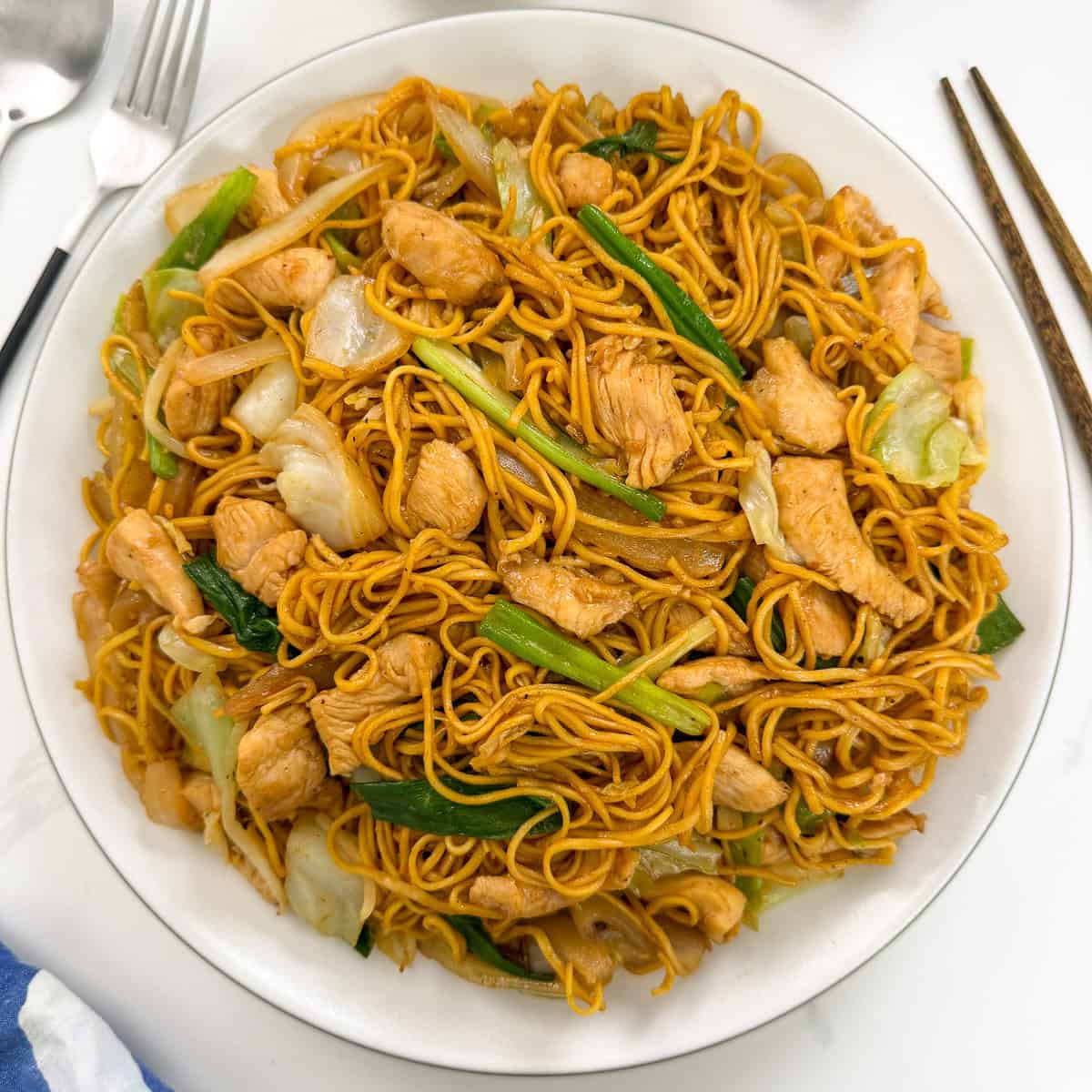A plate of Chicken Chow Mein featuring stir-fried noodles with tender chicken pieces, cabbage, and green onions. A fork, spoon, and chopsticks are placed nearby on a white surface.