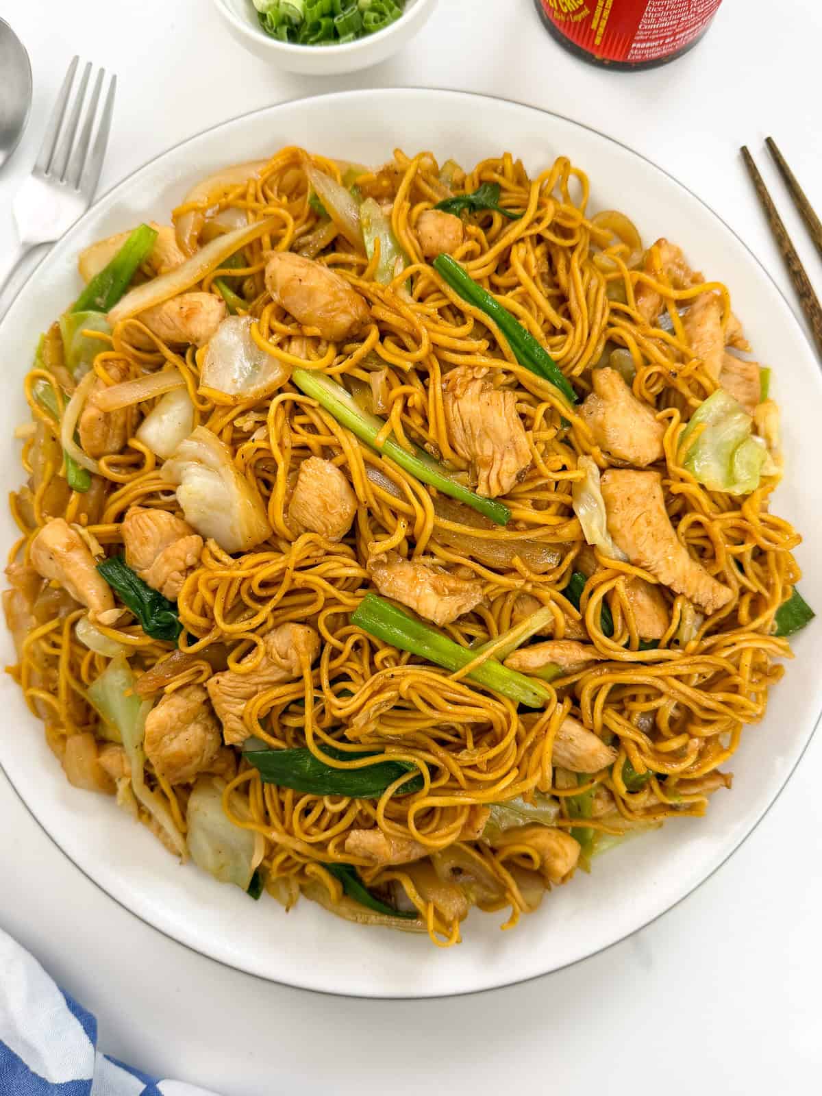 A plate of Chicken Chow Mein sits on a white dish, featuring stir-fried noodles with pieces of chicken, green onions, and cabbage. Nearby, a spoon, chopsticks, a small bowl of sliced green onions, and a bottle of sauce complete the inviting setting.
