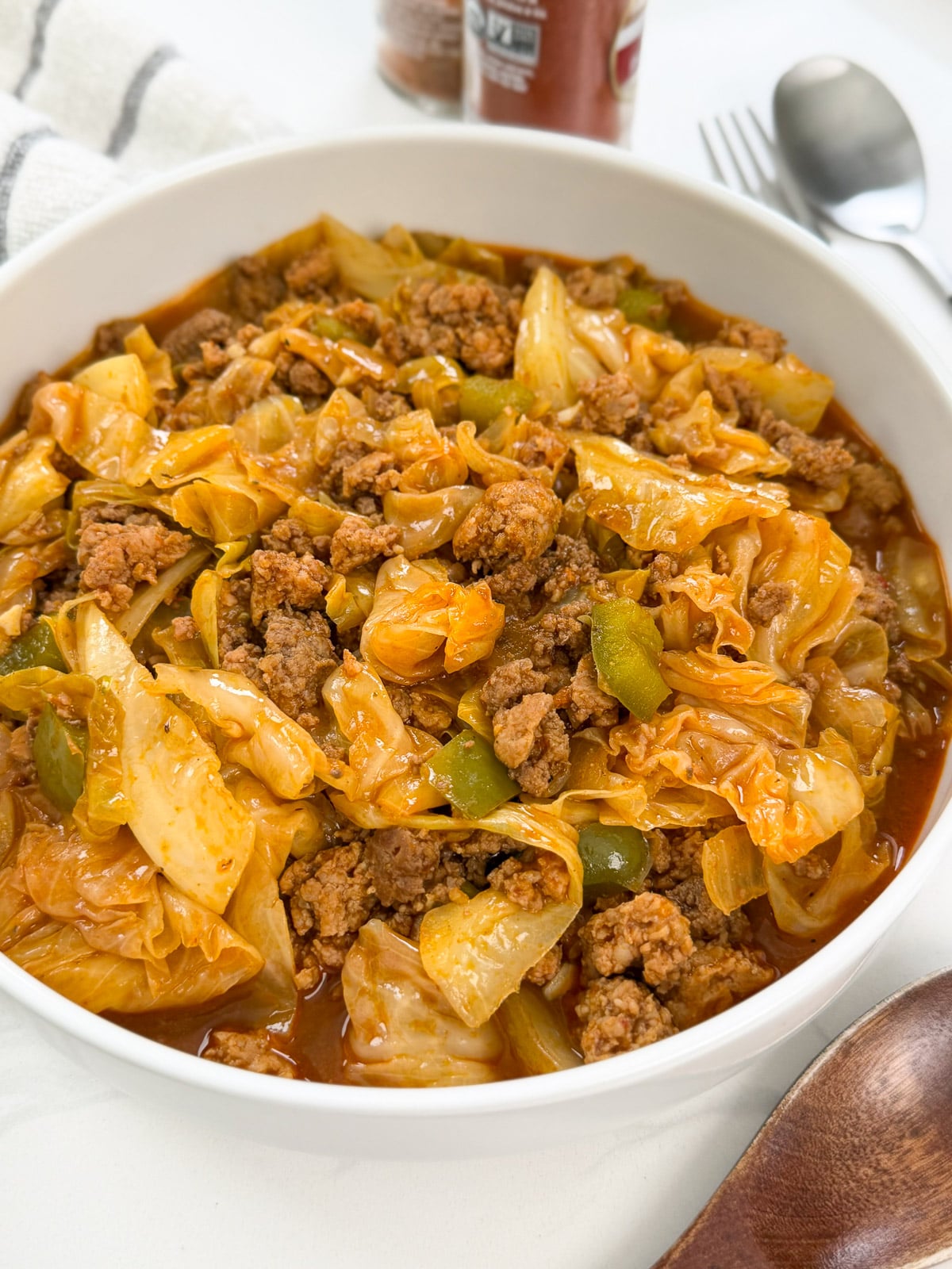 A white bowl filled with an unstuffed cabbage roll dish, also known as a Dirty Cabbage Recipe, featuring cooked ground beef, chopped cabbage, green peppers, and tomato sauce. A spoon, fork, and napkin are in the background.