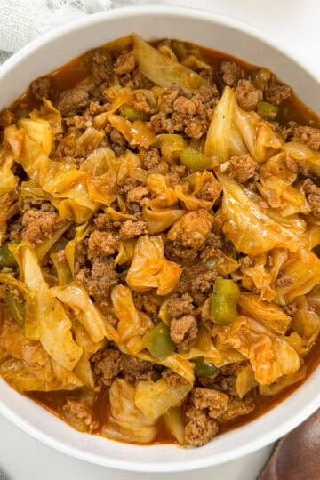 A white bowl filled with a cooked mixture of ground beef, chopped cabbage, and green bell peppers in a red sauce—this irresistible dish, known as Viral Dirty Cabbage, is placed on a white surface next to utensils and a wooden spoon.