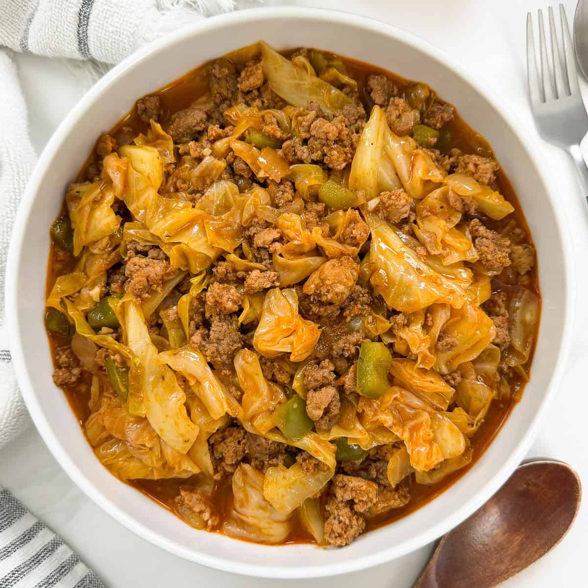 A white bowl filled with a cooked mixture of ground beef, chopped cabbage, and green bell peppers in a red sauce—this irresistible dish, known as Viral Dirty Cabbage, is placed on a white surface next to utensils and a wooden spoon.