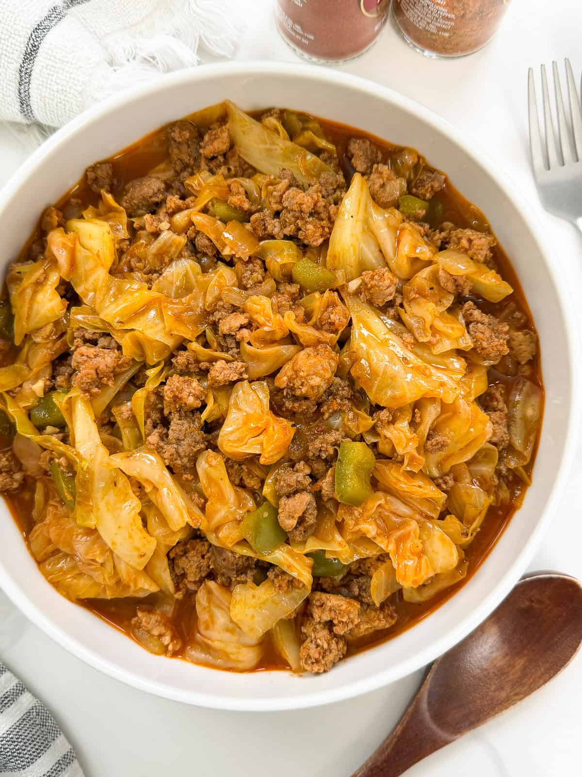 A white bowl filled with cooked cabbage, ground meat, and green bell peppers in a tomato-based sauce showcases the irresistible flavors of Viral Dirty Cabbage, with a wooden spoon and fork beside it on a white surface.