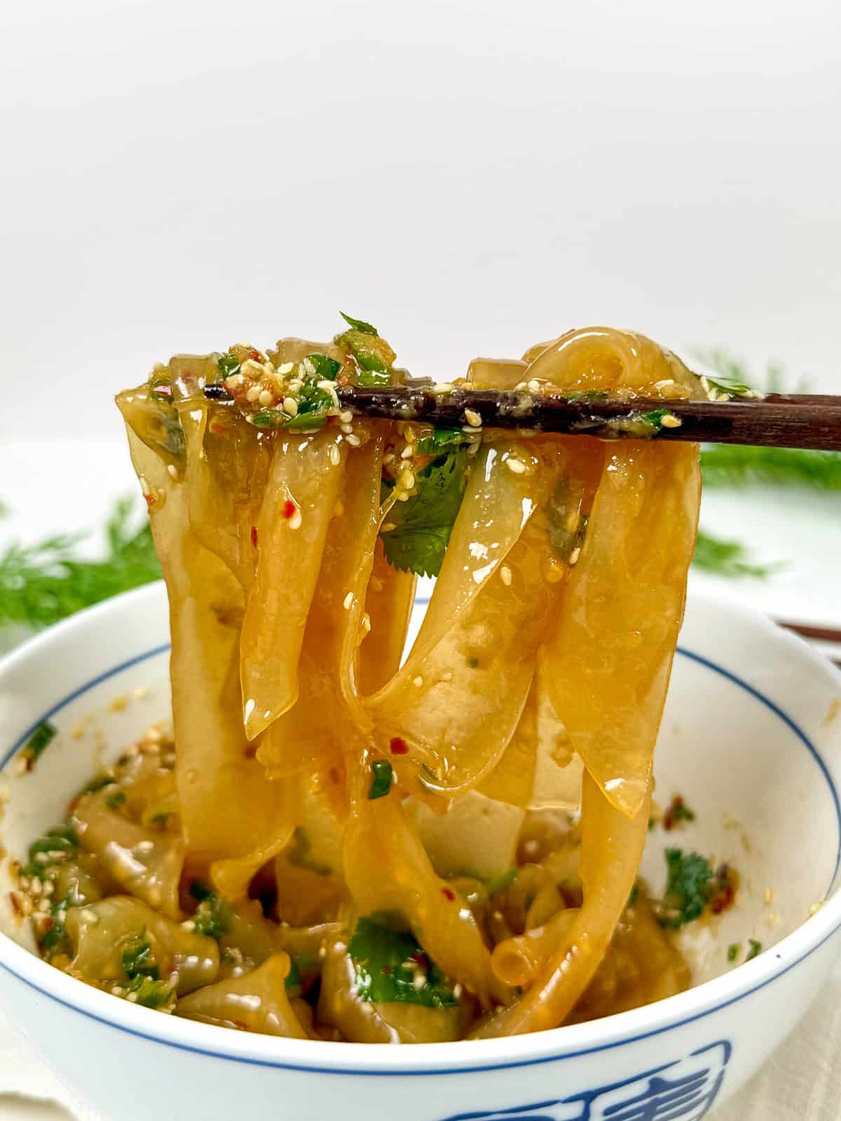 Chopsticks lifting wide, flat rice paper noodles coated in a spicy sauce with herbs, ginger scallion oil, and sesame seeds from a white bowl with blue rim.