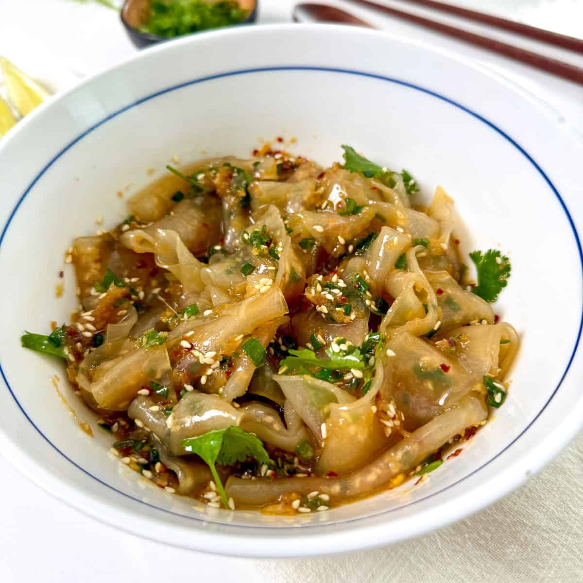 A white bowl filled with spicy chili oil dumplings garnished with sesame seeds, chopped cilantro, and red pepper flakes. Chopsticks and a small dish of ginger scallion oil are visible in the background, adding an aromatic flair to the presentation.