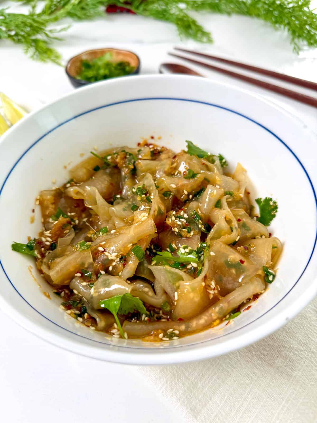 A white bowl filled with dumplings topped with chopped cilantro, sesame seeds, and chili flakes. Ginger scallion oil drizzles over the top, adding a fragrant touch. Chopsticks and a small dish of green herbs are in the background.