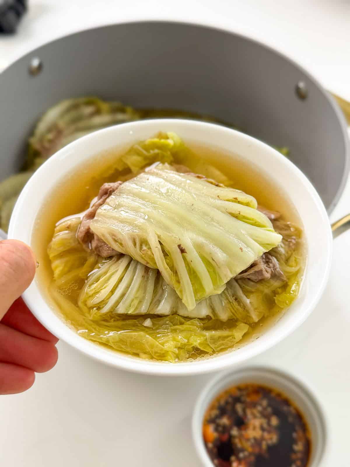 A bowl of cabbage-wrapped meat in clear broth, held above a pot with more of the same Mille-Feuille Nabe (Japanese Hot Pot) dish. A small bowl of dipping sauce with chili and garlic is nearby.