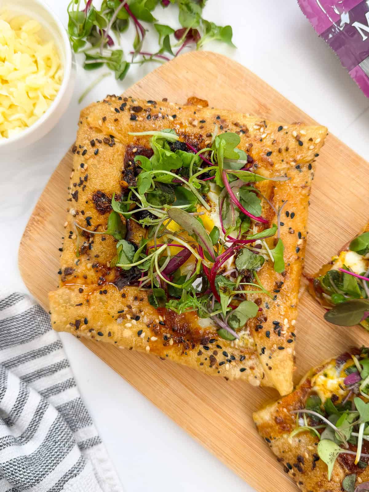 A savory galette topped with fresh microgreens and black sesame seeds sits on a wooden board, reminiscent of a Rice Paper Breakfast Danish, with a bowl of shredded cheese and a striped cloth nearby.