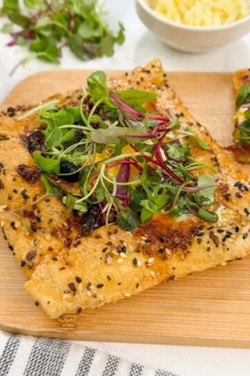 A square piece of seeded pastry, reminiscent of a Rice Paper Breakfast Danish, is topped with melted cheese, leafy greens, and microgreens as it sits on a wooden board next to another slice and a bowl of grated cheese.
