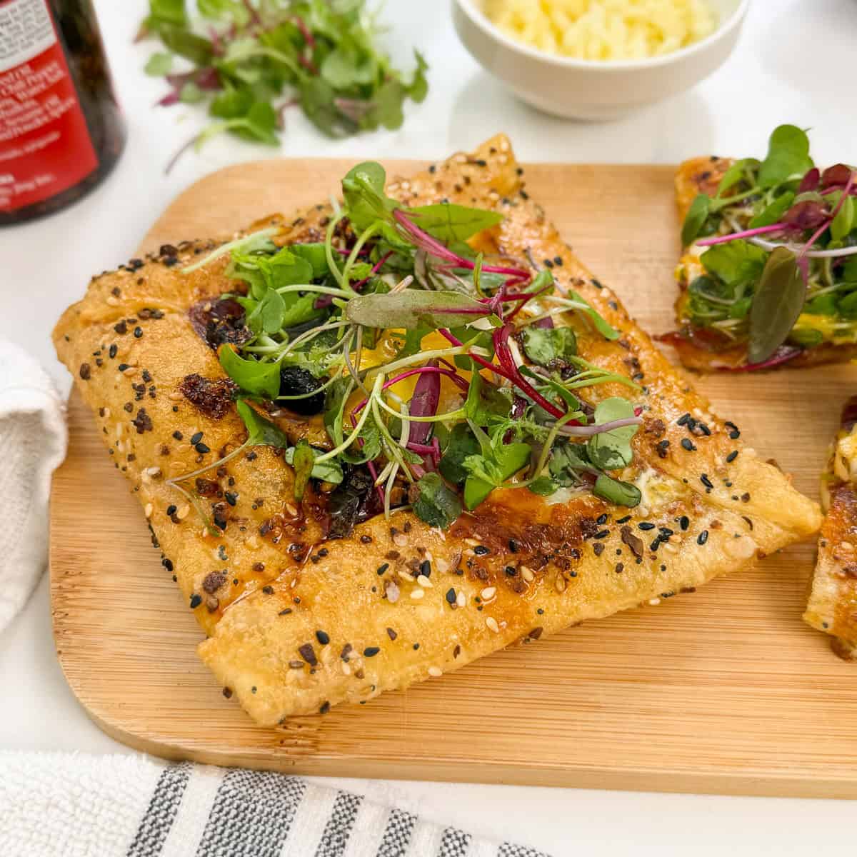 A square piece of seeded pastry, reminiscent of a Rice Paper Breakfast Danish, is topped with melted cheese, leafy greens, and microgreens as it sits on a wooden board next to another slice and a bowl of grated cheese.