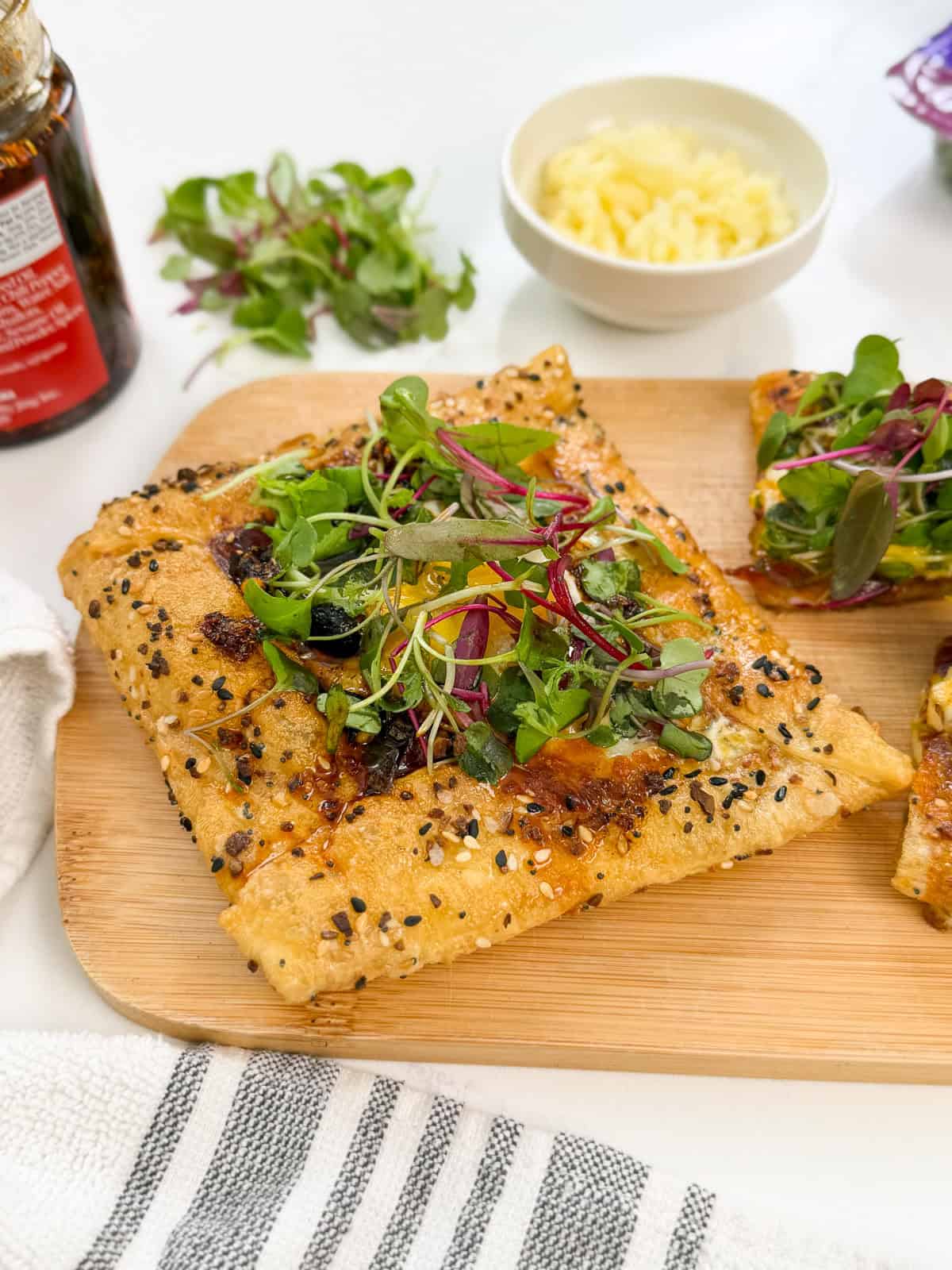Square piece of flatbread—reminiscent of a Rice Paper Breakfast Danish—topped with melted cheese, fresh greens, and seeds on a wooden board, with a bowl of shredded cheese and a bottle in the background.