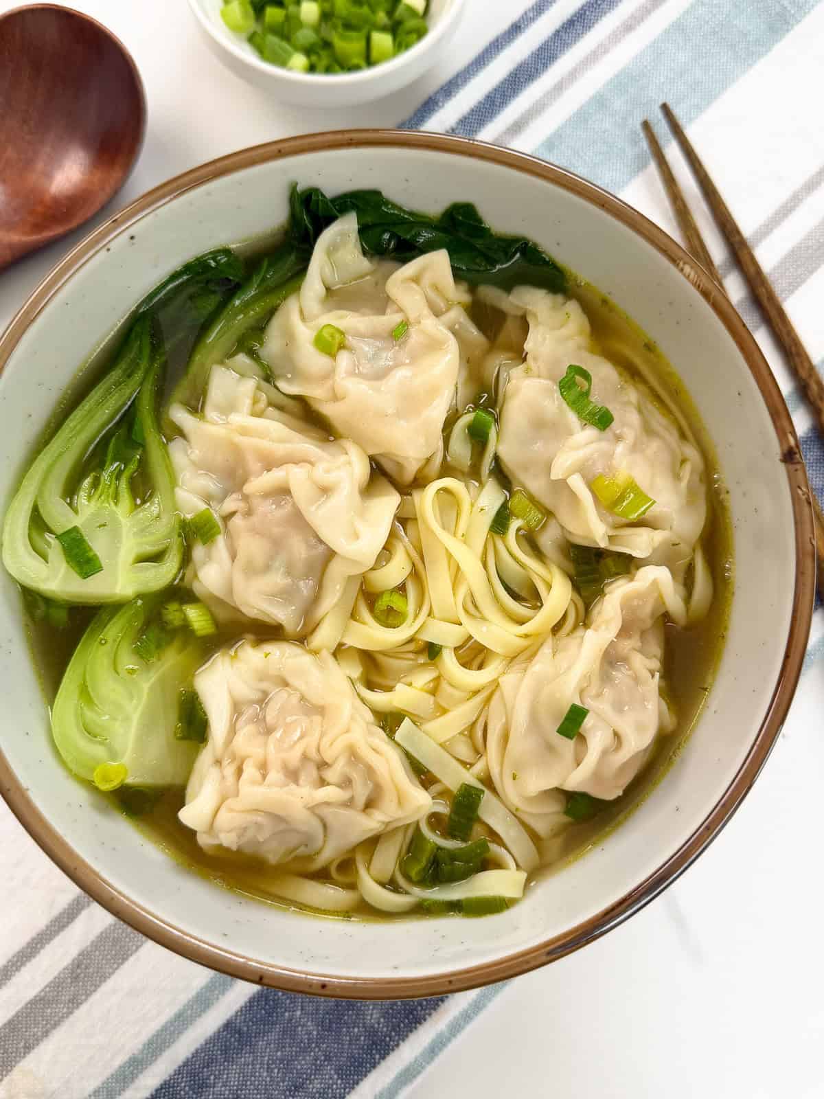 A steaming bowl of Wonton Noodle Soup with delicate dumplings, crisp bok choy, and fresh green onions sits on a striped cloth, accompanied by a pair of chopsticks.