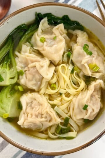 Bowl of wonton noodle soup with dumplings, bok choy, sliced green onions, and noodles in broth. Wooden spoon and chopsticks are next to the bowl on a striped cloth.