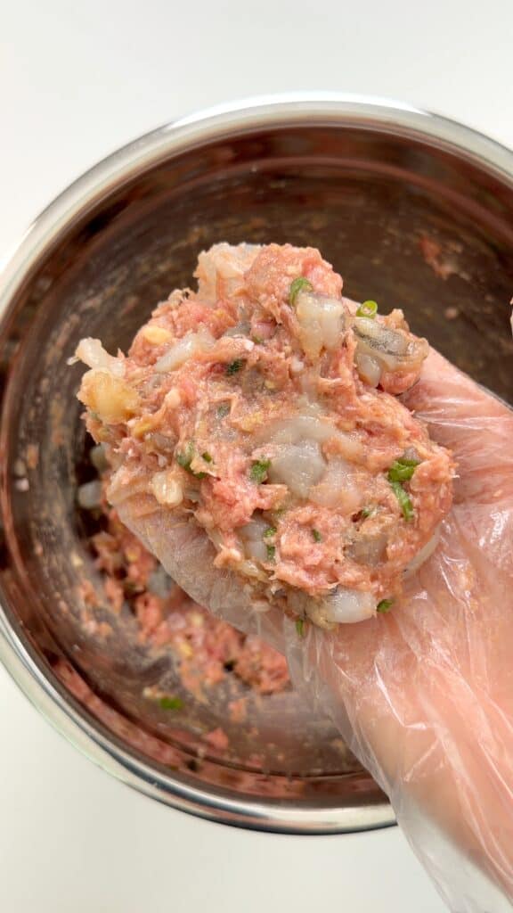 A gloved hand holds a mixture of ground meat, shrimp, and herbs over a metal bowl filled with the same mixture, ready to be shaped into patties or balls.