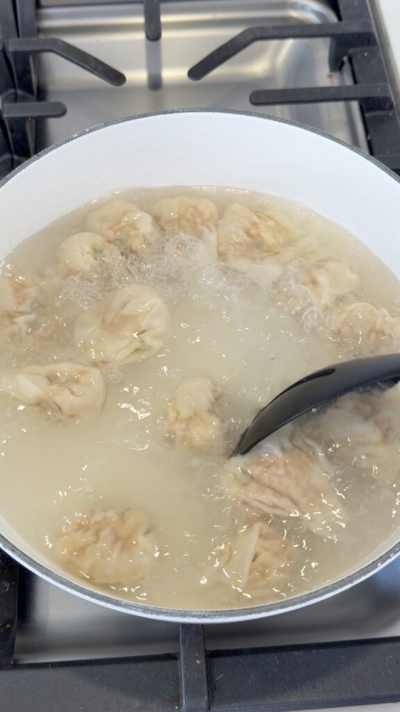 A white pot on a stove with dumplings boiling in cloudy water. A black ladle is stirring the dumplings as they cook.