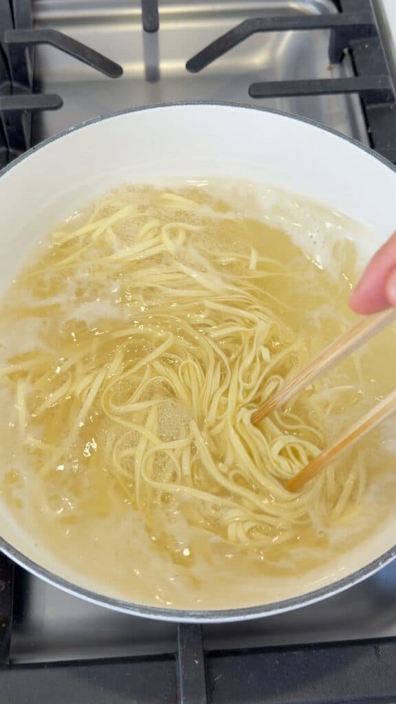 A hand uses chopsticks to stir noodles boiling in a pot of water on a stovetop.