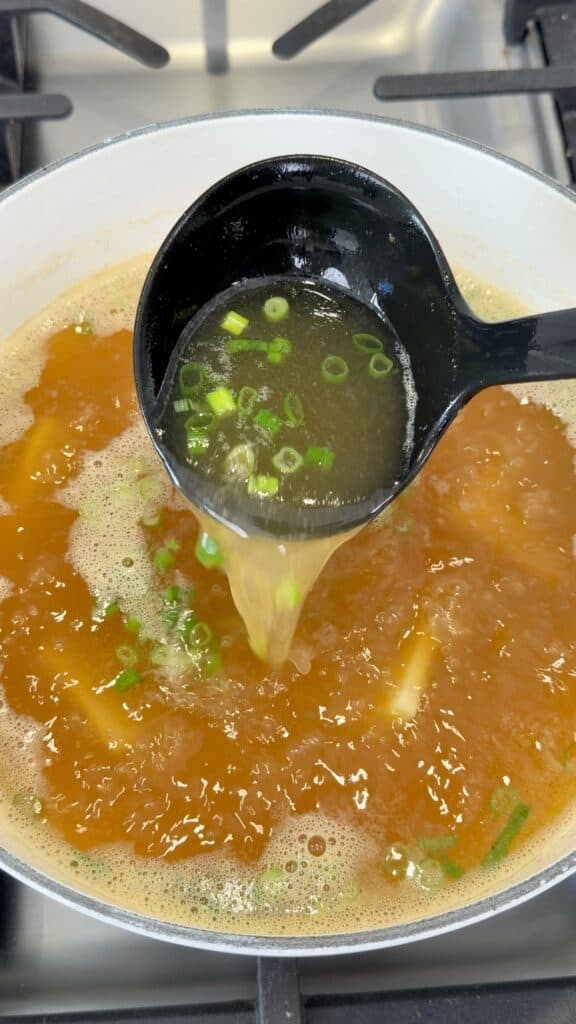 A black ladle pours broth with chopped green onions into a pot of simmering soup on a stovetop.
