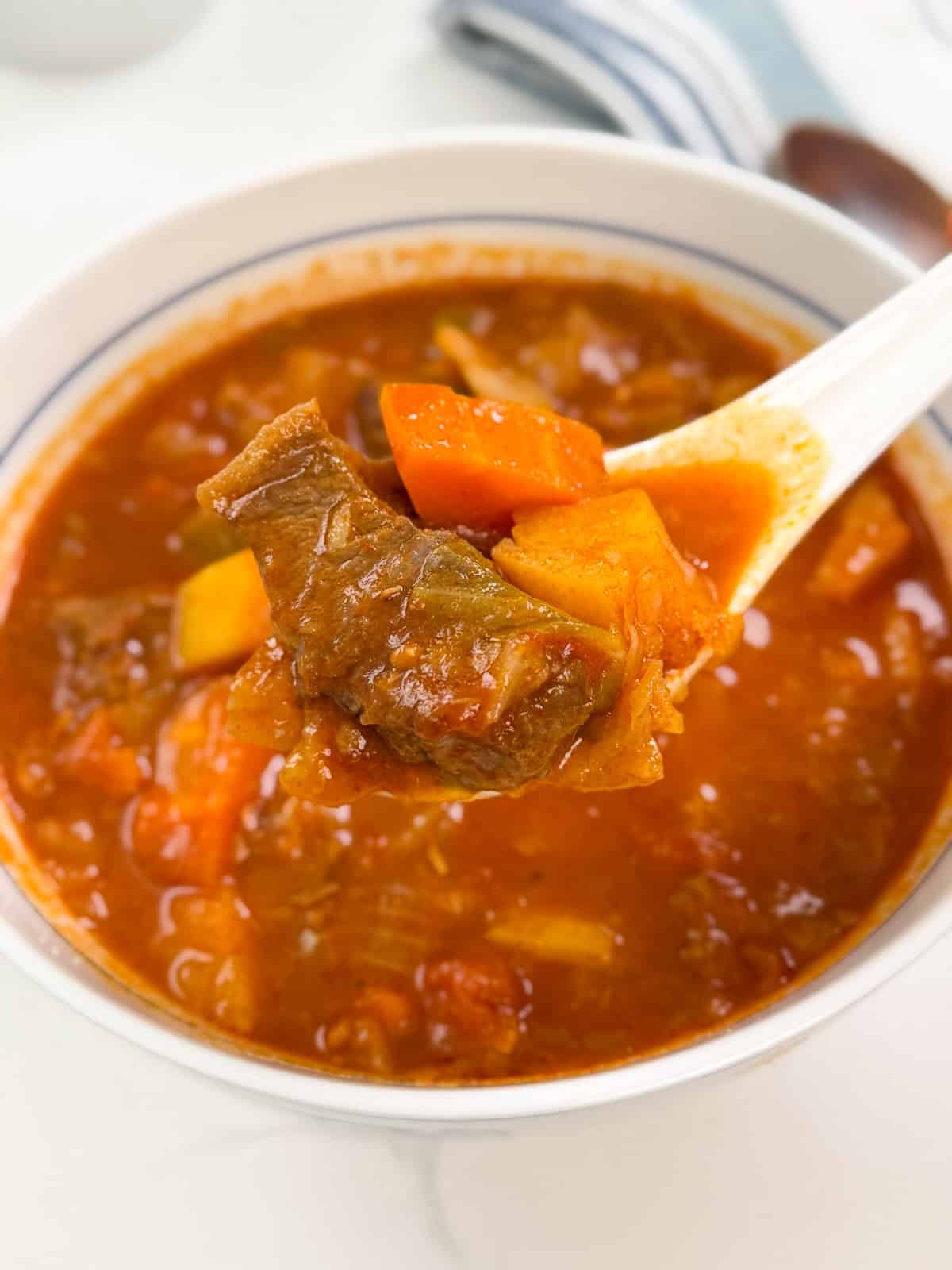 A white spoon lifting a piece of beef, potato, and carrot from a bowl of vegetable beef soup.
