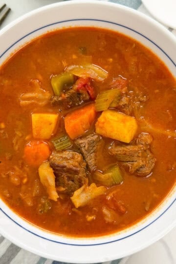 A white bowl of beef vegetable soup containing chunks of beef, carrots, celery, and potatoes in a tomato-based broth.