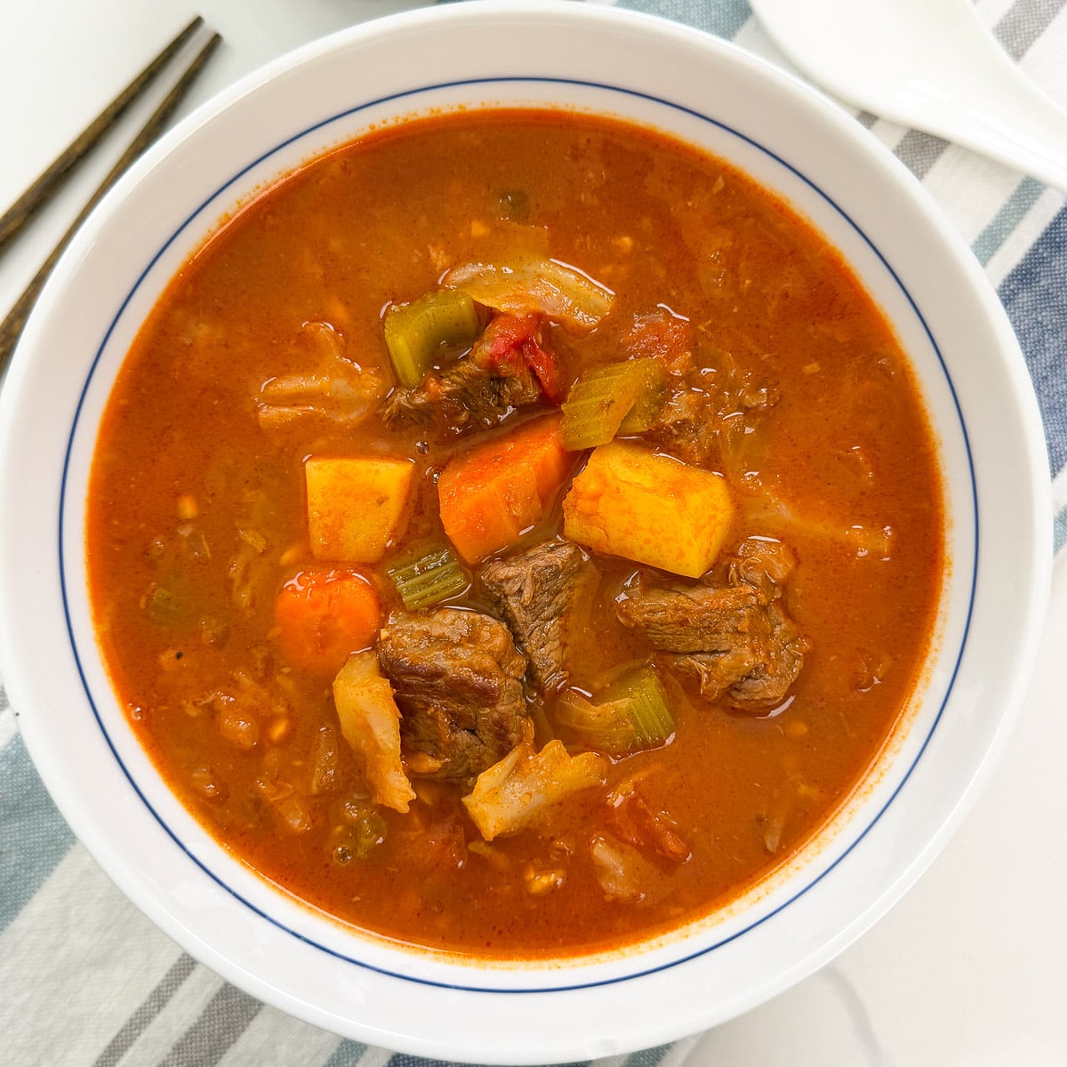 A white bowl of beef vegetable soup containing chunks of beef, carrots, celery, and potatoes in a tomato-based broth.