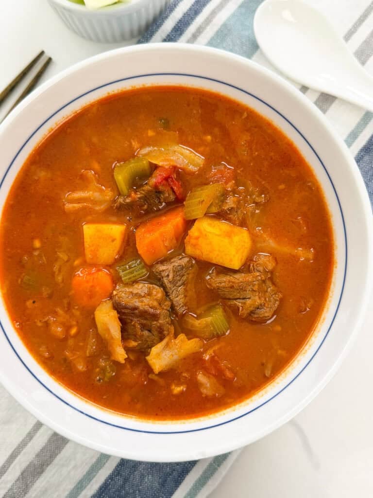A bowl of beef stew with chunks of meat, carrots, and celery in a red tomato-based broth.