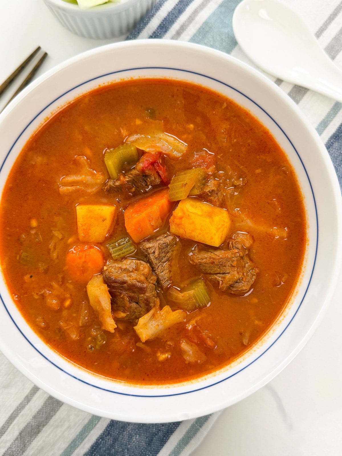 A bowl of beef stew with chunks of meat, carrots, and celery in a red tomato-based broth.