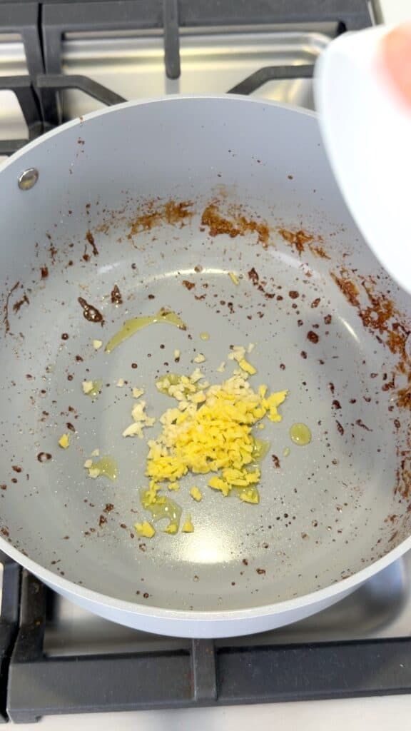 Minced garlic and ginger being added to a pan with browned bits and oil.
