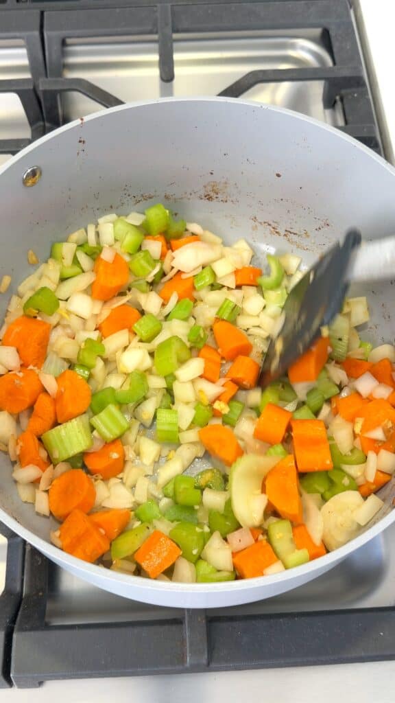A spatula stirs chopped carrots, celery, and onions in a light gray pan on a stovetop.