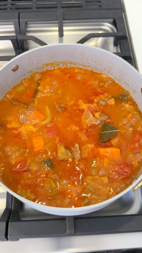 A light-colored pot on a stovetop filled with a simmering vegetable and beef stew featuring a visible bay leaf.
