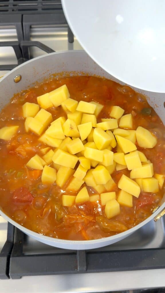 Diced raw potatoes being poured from a white bowl into a pan of simmering tomato-based stew.