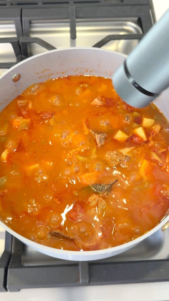 A hand grinds black pepper over a bubbling pot of vegetable and beef stew on a stovetop.