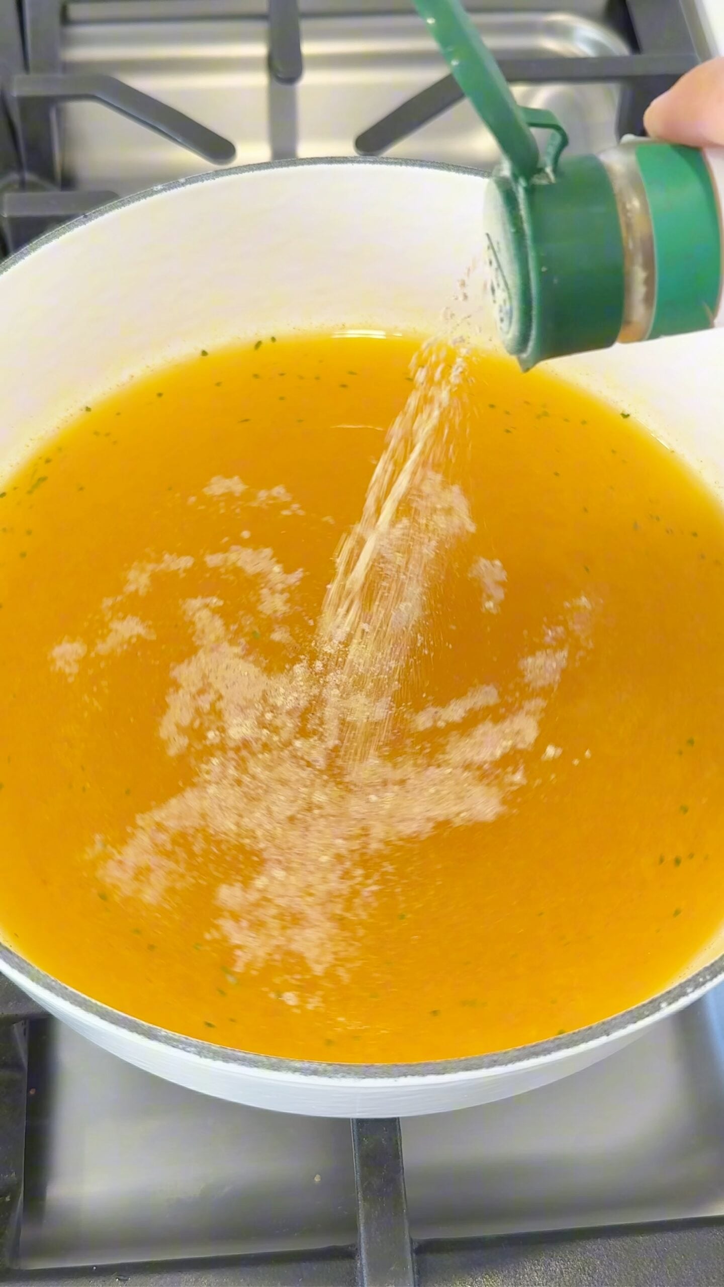 A hand sprinkles seasoning from a green shaker into a pot of broth on a stove.