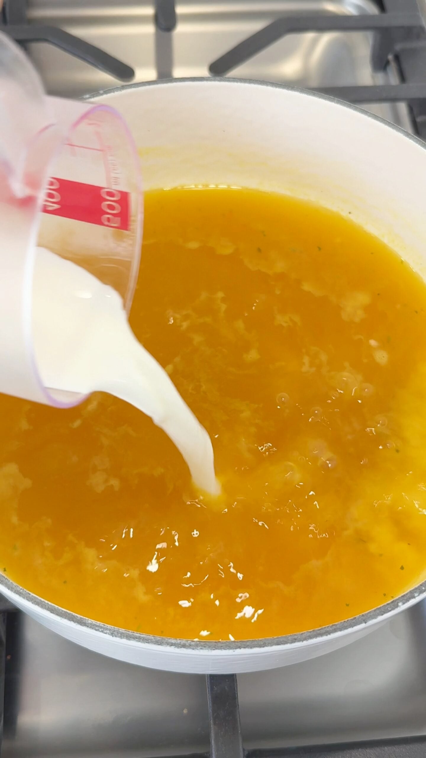 A measuring cup pours milk into a pot of orange soup on a stovetop.