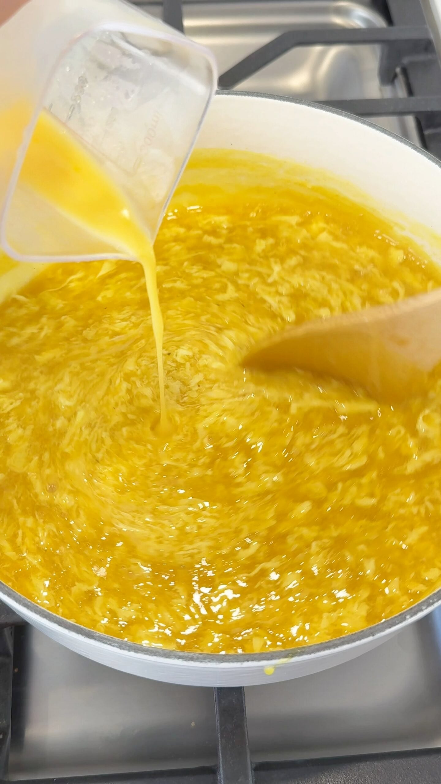 Egg mixture is being poured into a pot of hot liquid, with a wooden spoon stirring the contents, likely making egg drop soup on a stovetop.