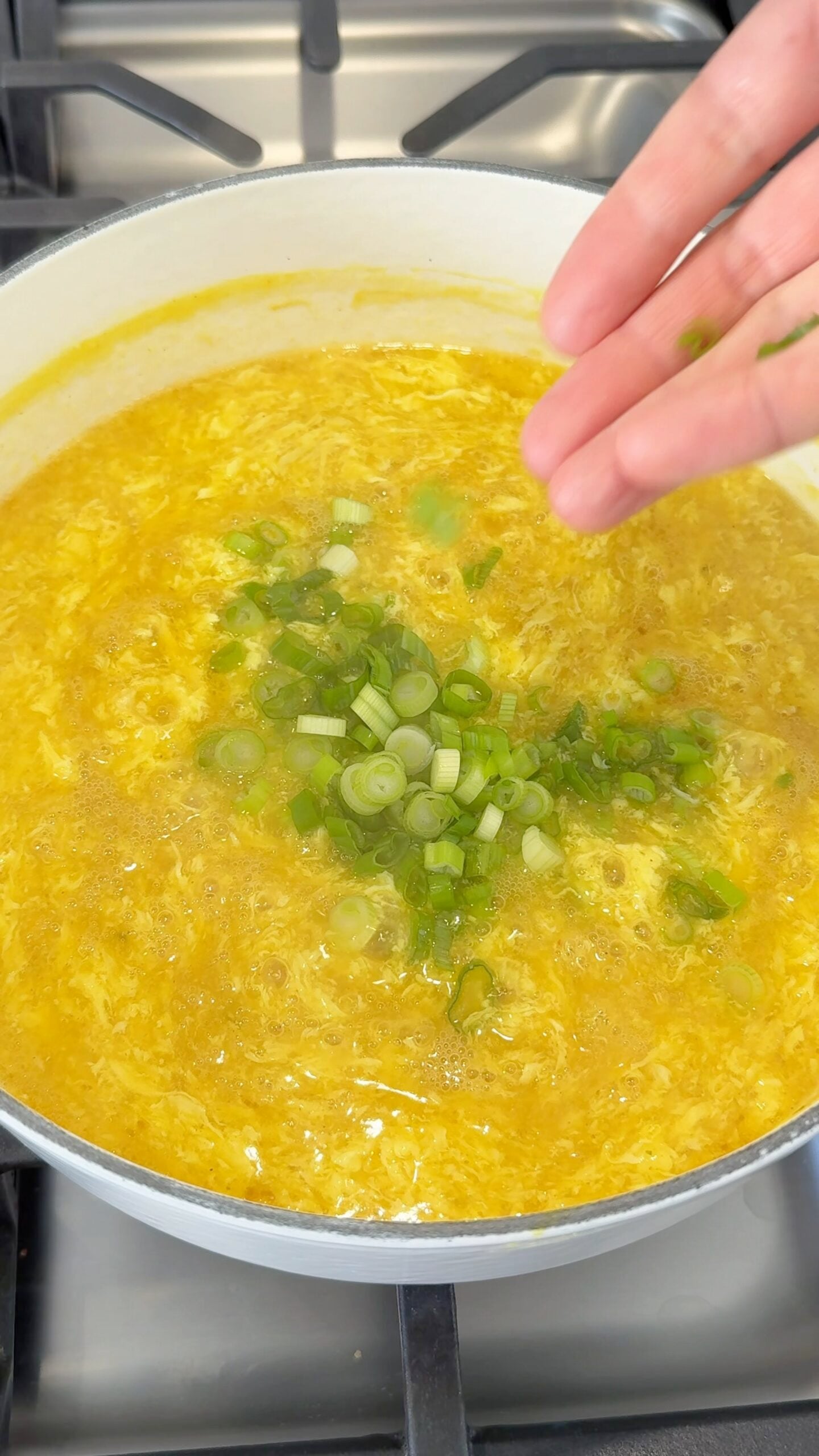A hand sprinkles chopped green onions into a pot of yellow soup simmering on a stovetop.