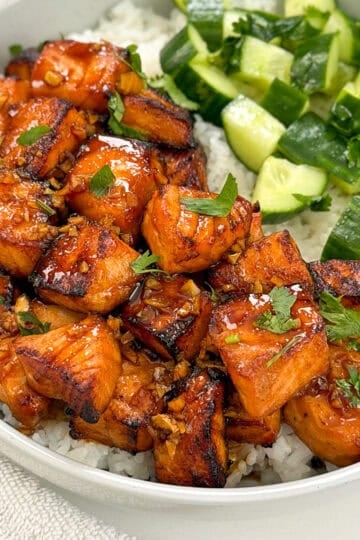 A bowl of white rice topped with glazed salmon bites and diced cucumber, garnished with fresh herbs.